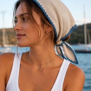 May include: A beige knit headscarf with a blue trim, tied at the back. The headscarf is worn over the head and tied at the nape of the neck. A gold necklace with small beads is also visible.
