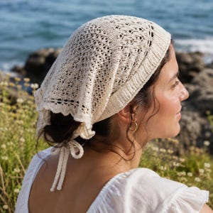 Könnte beinhalten: Eine cremefarbene Strickhaube mit gewelltem Rand und Bindeband. Die Haube hat ein Lochmuster. Der Hintergrund zeigt das Meer und Wildblumen. Getragen von einer Person mit dunklem Haar.