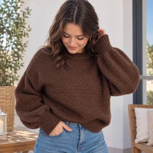 Könnte beinhalten: Ein dunkelbrauner, langärmeliger Strickpullover mit Rundhalsausschnitt und gerippten Bündchen. Der Pullover hat ein grobes, strukturiertes Strickmuster mit überschnittenen Schultern und einer lockeren Passform, kombiniert mit einer hellblauen Jeans.