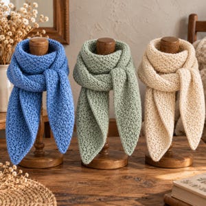 May include: Three knitted scarves in blue, sage green, and cream, each featuring a textured stitch pattern. The scarves are draped over individual wooden stands, displaying their triangular shape and soft, woven construction.