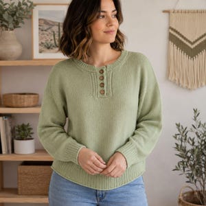 May include: A sage green long-sleeve knit jumper featuring a round neckline with a short button placket containing four small brown buttons. The jumper has ribbed cuffs and a ribbed hem, with a smooth, solid-coloured knit texture throughout.