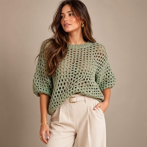 Puede incluir: Un suéter de ganchillo color verde salvia con un patrón de malla abierta y holgada, cuello redondo y mangas abullonadas hasta el codo. La prenda se combina con pantalones beige de talle alto que incluyen un cinturón a juego con hebilla dorada.