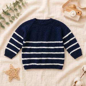 Peut inclure: Un pull en tricot à manches longues présentant une base bleu marine foncé avec des rayures horizontales blanches sur le corps et les manches. Le vêtement possède une encolure ronde et des bords-côtes aux poignets et à la base.