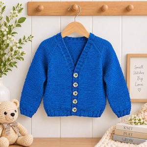 May include: A vibrant royal blue long-sleeve knit cardigan featuring a V-neckline, ribbed cuffs and hem, and five round buttons with a colorful floral pattern. The garment is displayed on a wooden hanger against a white paneled wall.