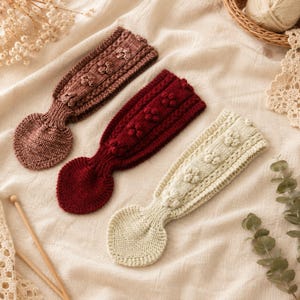 May include: Three knitted headbands in dusty rose, deep burgundy, and cream. Each headband features a textured pattern of small raised bobbles along the centre and a rounded, heart-shaped knit section at one end, showcasing detailed yarn work.
