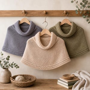 Op de afbeelding: Drie gebreide poncho's met colkraag hangend aan houten kledinghangers. De poncho's zijn in de kleuren stoffig blauw, beige en olijfgroen. Elk kledingstuk heeft een tricotsteek lijfje met een textuurrijke geribbelde rand langs de zoom en hals.