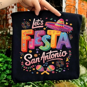 Puede incluir: Camiseta negra con la frase "Let's Fiesta San Antonio" en letras coloridas y decorativas. El diseño incluye un sombrero, maracas y notas musicales, celebrando un evento festivo.