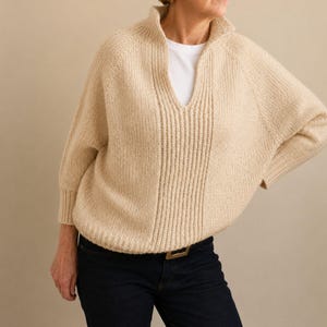 May include: A beige knit sweater featuring a high collar with a deep V-neck opening, raglan sleeves, and a vertical ribbed panel down the center front. The sweater is worn over a plain white t-shirt and paired with dark blue denim jeans.
