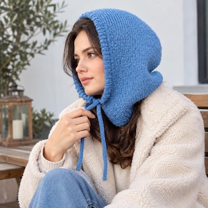 May include: A blue knitted hood with a ribbed edge and two long matching knitted ties hanging down. The hood is constructed from a textured yarn and features a rounded shape that covers the head, providing a snug fit around the face and chin.