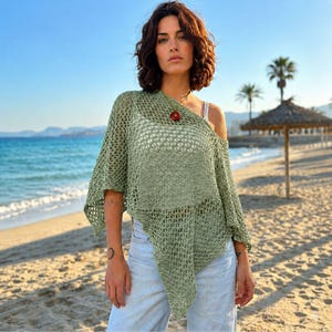 May include: A sage green, loose-fitting, open-knit crochet poncho top with an asymmetrical hemline, worn over a white camisole, paired with light-wash denim jeans and a circular red pendant necklace on a thin cord.
