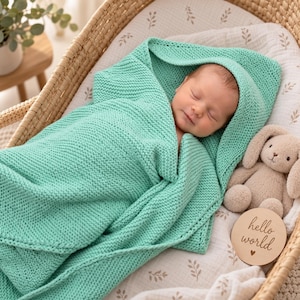 Puede incluir: Una manta de bebé tejida en color verde menta envuelve a un bebé dormido. La manta presenta un patrón de punto bobo texturizado. Junto al bebé hay un conejo de peluche beige y un disco de madera con el texto hello world grabado.
