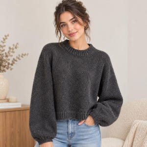 Puede incluir: Un suéter de manga larga en color gris carbón con cuello redondo y acabado de punto texturizado. La prenda presenta hombros caídos, mangas voluminosas con puños acanalados y un dobladillo acanalado, combinado con pantalones vaqueros de color azul claro.