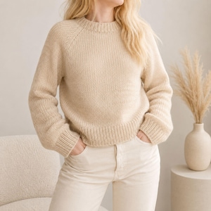 May include: A beige long-sleeved sweater featuring a crew neckline and a ribbed knit pattern at the collar, cuffs, and hem. The garment is constructed with a stockinette stitch throughout, providing a consistent texture across the torso and sleeves.
