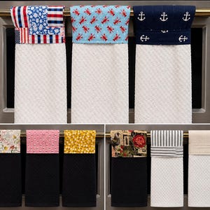 May include: A collection of kitchen towels with various patterned tops. Designs include an American flag, lobsters on blue, anchors on navy, floral, pink polka dots, yellow flowers, a rooster, stripes, and solid beige. The towels have a white or black textured base.