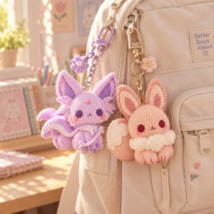 May include: Two crocheted animal keychains attached to a beige backpack. One is a lavender fox-like creature with purple accents and a flower charm. The other is a peach-colored rabbit-like figure with a white chest and a small flower charm.