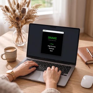 May include: A laptop screen displaying the classic retro game "Snake". The menu shows options to play, view high scores, adjust settings, or quit. A person is using the laptop. A cup of coffee, a notebook, and a mouse are on the wooden desk.