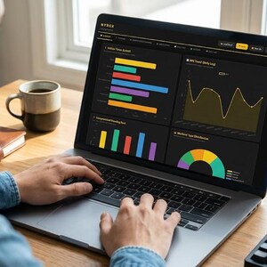 May include: A person using a laptop displaying data visualizations. The screen shows various charts and graphs in multiple colors, including bar graphs, line graphs, and a pie chart. A coffee mug, notebook, and keyboard are also visible on the desk.
