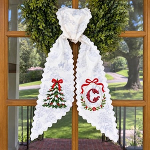 Puede incluir: Un colgador de puerta de tela blanca con estampado toile azul, bordes festoneados y dos extremos largos. Un extremo muestra un árbol de Navidad bordado en verde con un lazo rojo, el otro una letra C roja en una corona.