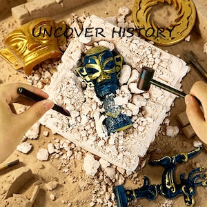 May include: An archaeological dig kit with the text "UNCOVER HISTORY". The kit includes a blue and gold mask, a golden tiki-like figure, and a hammer. The scene is set on a sandy surface with fragments of a brick-like structure.