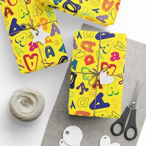 May include: Wrapped gifts with yellow paper featuring colourful letter designs. Each gift is tied with twine and a white tag. A ball of twine, scissors, and blank tags are also visible.