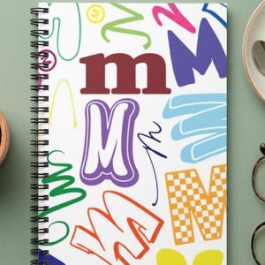 May include: A spiral-bound notebook with a white cover featuring a repeating pattern of the letter M in various fonts, colors, and styles, including solid red, purple with white outlines, orange outlines, and an orange and white checkered pattern.
