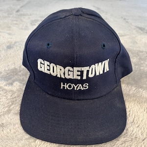 May include: A navy blue baseball cap with a flat brim. The cap features the white embroidered text "GEORGETOWN" above "HOYAS". The cap has two small air vents on the crown.