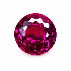 Puede incluir: Una piedra preciosa redonda y facetada de un color rojo magenta profundo y vibrante. La piedra presenta un corte brillante con numerosas facetas triangulares y en forma de cometa que reflejan la luz, creando un aspecto brillante y claro.