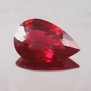 May include: A faceted, pear-shaped ruby gemstone. The gem is a deep, vibrant red color with multiple facets that reflect light. The ruby is displayed on a reflective surface, creating a mirrored effect. The overall shape is elongated with a rounded top and a pointed bottom.