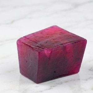 May include: A rectangular block of translucent soap with a deep magenta and dark purple marbled color pattern. The surface of the soap has a slightly uneven, textured appearance with visible variations in color density throughout the solid form.