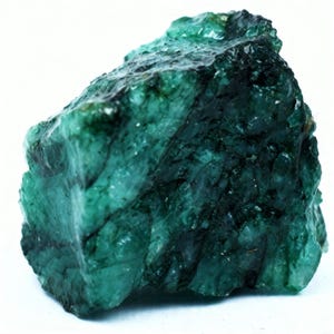 Puede incluir: Una piedra preciosa de esmeralda en bruto y sin pulir con un color verde intenso y una textura cristalina irregular. La piedra presenta variaciones naturales en la opacidad e inclusiones oscuras en toda su superficie, mostrando una forma rugosa.