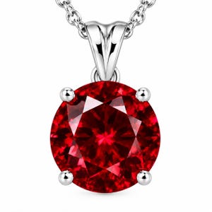 May include: A round, faceted, deep red gemstone pendant set in a four-prong silver-toned metal setting. The pendant is attached to a silver-toned chain link necklace, featuring a polished finish and a classic solitaire design.