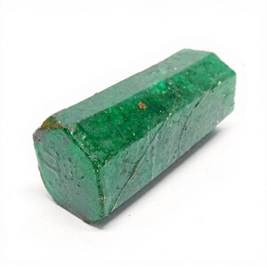 Puede incluir: Un cristal de esmeralda en bruto de forma hexagonal, con un color verde intenso y una superficie texturizada semitransl&uacute;cida. La piedra presenta facetas naturales, inclusiones internas visibles y un peque&ntilde;o dep&oacute;sito mineral marr&oacute;n rojizo.