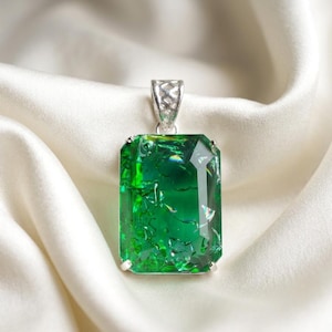 May include: A rectangular pendant featuring a large, faceted emerald green stone with internal reflective inclusions, held by four silver prongs and topped with a decorative silver filigree bail, showcasing a vibrant and clear green hue.
