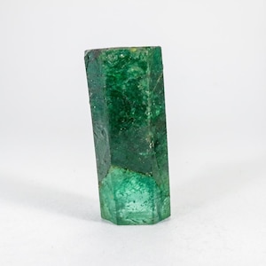 Puede incluir: Un cristal de esmeralda hexagonal en bruto que presenta un tono verde bosque profundo con inclusiones internas visibles y texturas superficiales naturales. La piedra preciosa muestra una estructura prism&aacute;tica vertical alargada.