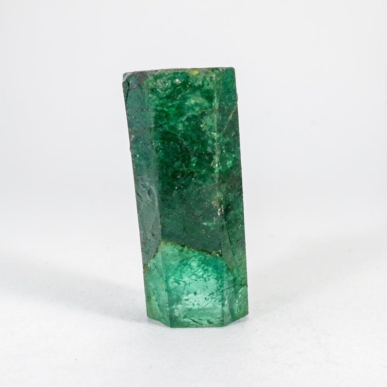 Puede incluir: Un cristal de esmeralda hexagonal en bruto que presenta un tono verde bosque profundo con inclusiones internas visibles y texturas superficiales naturales. La piedra preciosa muestra una estructura prism&aacute;tica vertical alargada.