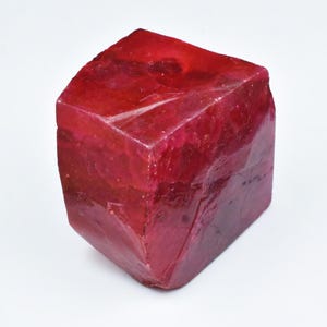 May include: A faceted, deep red gemstone with a cube-like shape. The surface of the stone has a rough, natural texture and a rich, saturated color. The gemstone appears to be a ruby, showcasing its natural beauty.