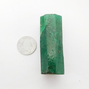 Puede incluir: Un cristal de esmeralda en bruto, alargado y hexagonal, de color verde intenso con una superficie natural texturizada. La piedra est&aacute; colocada junto a una moneda circular de color plateado para servir de escala visual.