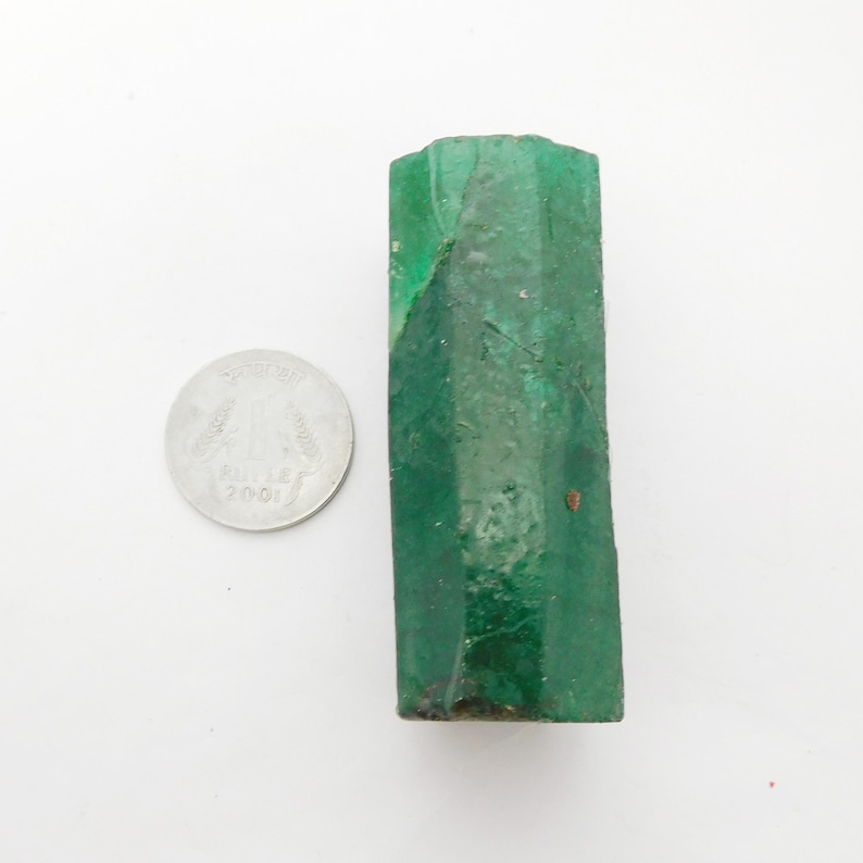 Puede incluir: Un cristal de esmeralda en bruto, alargado y hexagonal, de color verde intenso con una superficie natural texturizada. La piedra est&aacute; colocada junto a una moneda circular de color plateado para servir de escala visual.