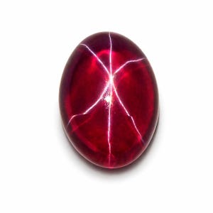 May include: An oval-shaped cabochon gemstone in a deep, translucent red colour. The stone features a distinct six-rayed star pattern, known as asterism, which appears as bright white intersecting lines across the smooth, polished surface.