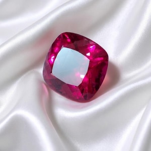Puede incluir: Una piedra preciosa de corte cojín con un tono magenta-rosado profundo y vibrante, con superficies facetadas que reflejan la luz. La piedra presenta una forma cuadrada con esquinas redondeadas y un aspecto pulido y translúcido.