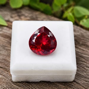 Può includere: Una gemma rosso scuro sfaccettata a forma di pera è appoggiata su un espositore quadrato bianco. Il taglio della gemma rivela molteplici sfaccettature, riflettendo la luce. L'insieme è su una superficie di legno, con fogliame verde sullo sfondo. È un rubino.