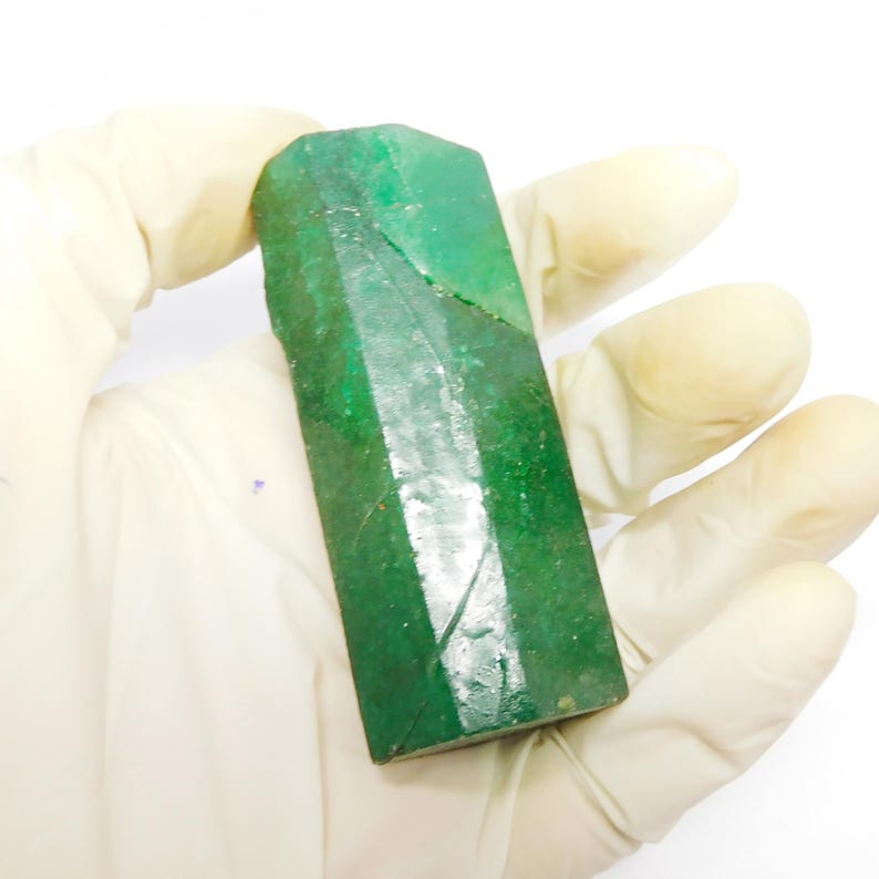 Puede incluir: Un cristal de esmeralda en bruto y alargado con un tono verde intenso y una estructura prism&aacute;tica hexagonal. La superficie muestra estr&iacute;as verticales naturales, inclusiones visibles y una calidad semitransl&uacute;cida con variados tonos de verde bosque.