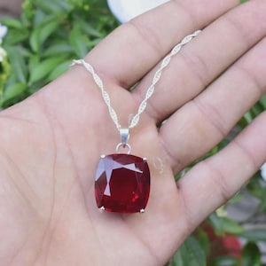 May include: A necklace featuring a large, cushion-cut, deep red gemstone pendant suspended from a delicate, twisted silver-tone chain. The faceted stone is secured by a simple metal bail, reflecting light across its polished, transparent surface.