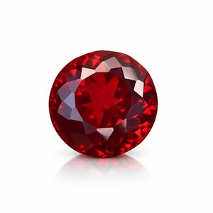 May include: A round, faceted, deep red gemstone. The gem is highly polished, reflecting light and creating a brilliant sparkle. The stone is set against a white background, with a subtle reflection below.