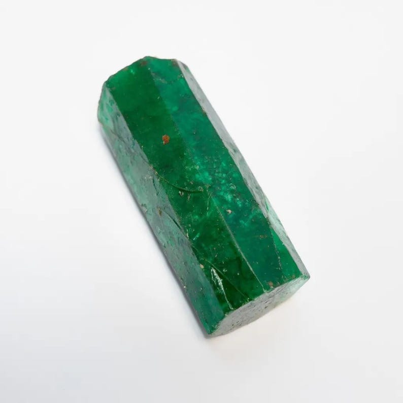 Puede incluir: Un cristal de esmeralda en bruto y alargado con forma de prisma hexagonal. La piedra muestra un color verde intenso y vibrante con fracturas internas visibles, inclusiones naturales y una textura semitransl&uacute;cida en toda su estructura.