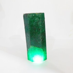 Puede incluir: Un esp&eacute;cimen de cristal de esmeralda en bruto de forma hexagonal con un color verde bosque profundo. La piedra presenta una superficie rugosa y texturizada con inclusiones naturales y un brillo transl&uacute;cido que emana desde la base iluminada.
