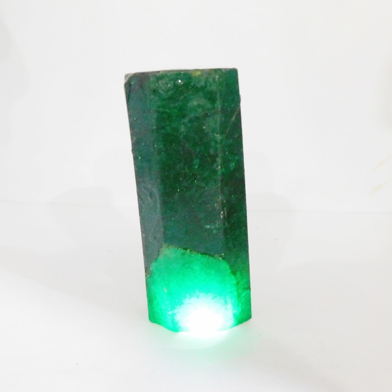 Puede incluir: Un esp&eacute;cimen de cristal de esmeralda en bruto de forma hexagonal con un color verde bosque profundo. La piedra presenta una superficie rugosa y texturizada con inclusiones naturales y un brillo transl&uacute;cido que emana desde la base iluminada.