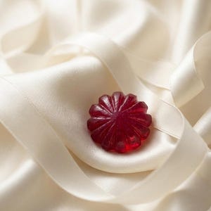 May include: A deep red, translucent, circular object with a fluted, star-like pattern radiating from the center. The object has a smooth, glossy finish and is resting within the folds of a soft, cream-colored, satin-like fabric with a sheen.