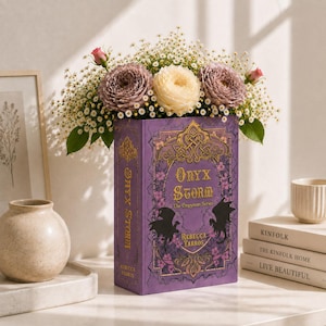 May include: A purple book titled Onyx Storm by Rebecca Yarros with gold filigree and black dragon silhouettes on the cover. The book is filled with a bouquet of white and mauve flowers, including ranunculus and baby's breath, emerging from the top.