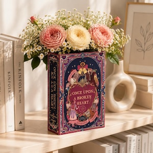 May include: A rectangular book-shaped vase featuring the title Once Upon a Broken Heart by Stephanie Garber on its dark blue and magenta cover, filled with a bouquet of pink and cream-colored ranunculus flowers and small white filler blossoms.